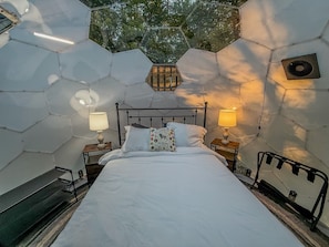 A room with a hexagonal ceiling, a bed with white bedding, two bedside lamps, and a window with a view of trees. - Leo | Black Lake Glamping Dome (Hammond)