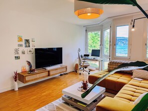 Ein modernes Wohnzimmer mit einem Flachbildfernseher, einem Computertisch, einer Couch und einem großen Fenster mit Blick auf Grünflächen und Gewässer.