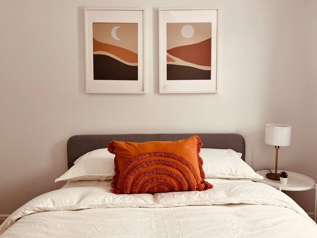 A neatly made bed with white linens and a single orange decorative pillow. There are two framed artworks above the bed, and a bedside table with a lamp to the right.