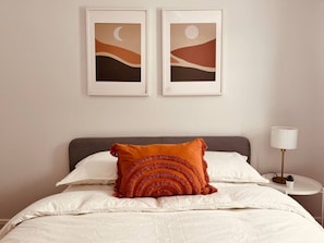 A neatly made bed with white linens and a single orange decorative pillow. There are two framed artworks above the bed, and a bedside table with a lamp to the right. - Modern apartment in the heart of Gatineau. (Gatineau)