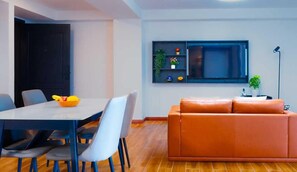 Una sala de estar moderna con un sofá, una mesa de comedor con sillas, un televisor montado en la pared y un pequeño estante con plantas.