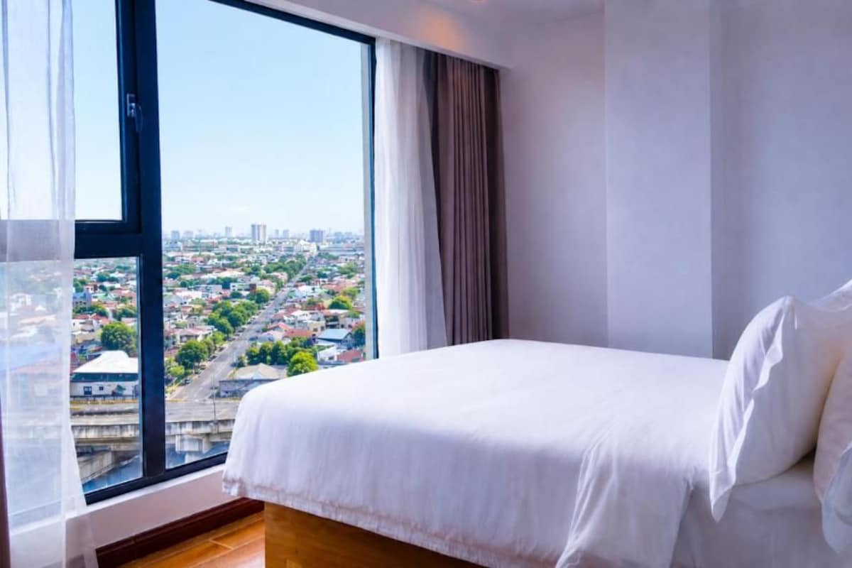 A hotel room with a large bed, a view of a cityscape through a window, and a curtain.