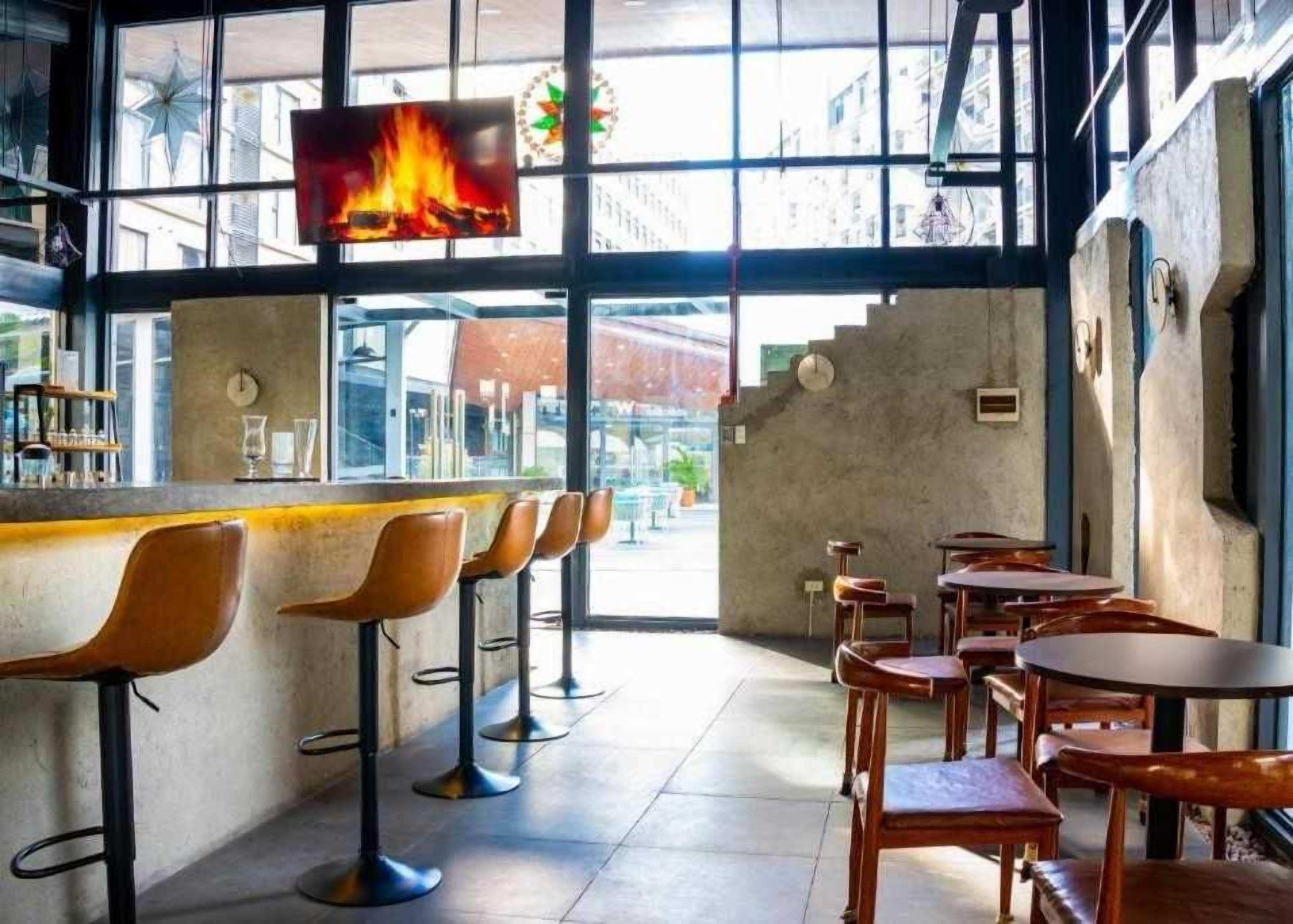 Un espace bar avec des tabourets et des tables, un écran de télévision diffusant une image de feu, et une vue sur la rue.