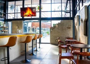 Zona de bar con taburetes y mesas, una pantalla de televisión mostrando un fuego, y vista de la calle.