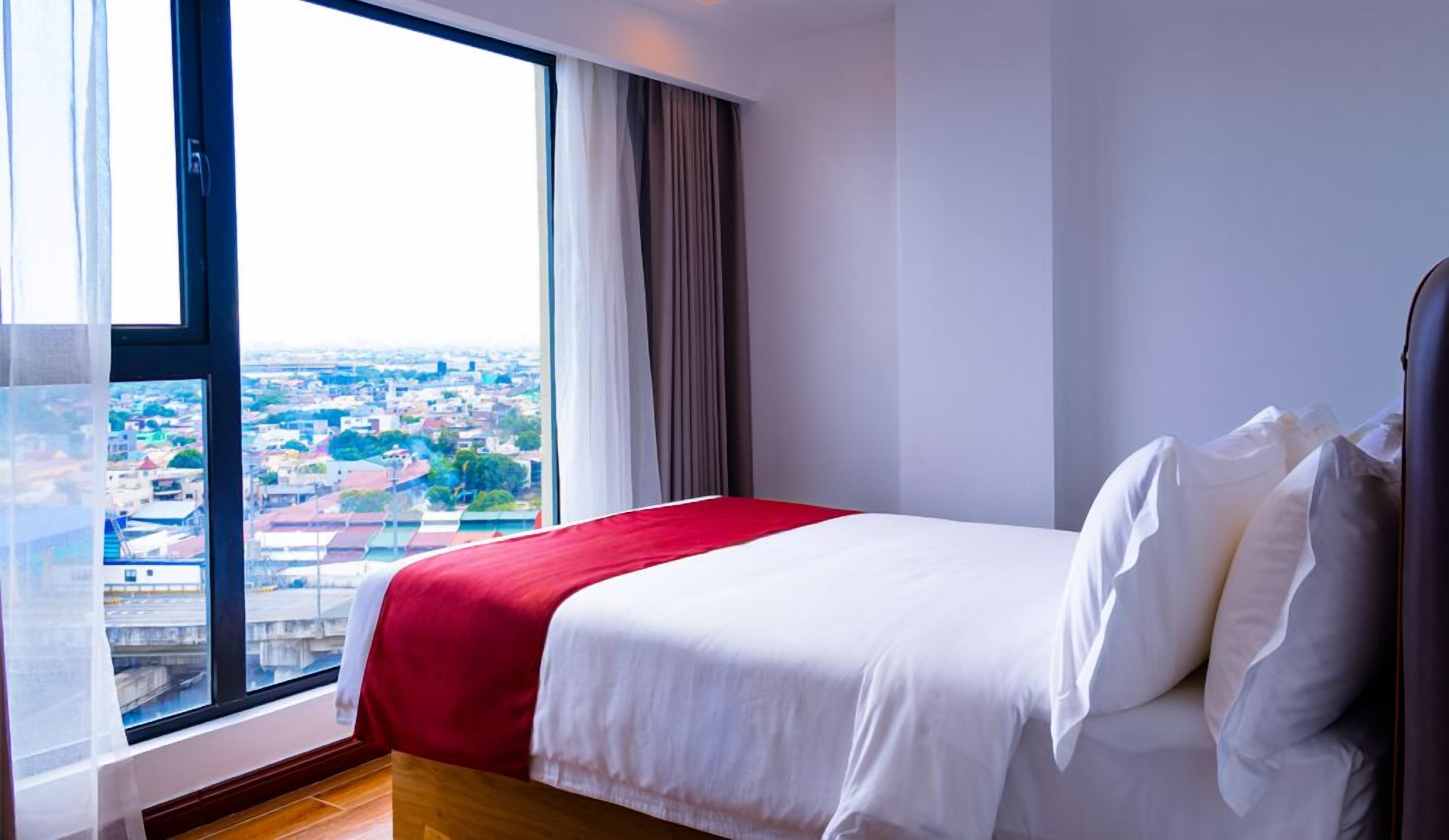 Quarto de hotel com uma grande janela, cama com roupa de cama branca e vista para a paisagem urbana.