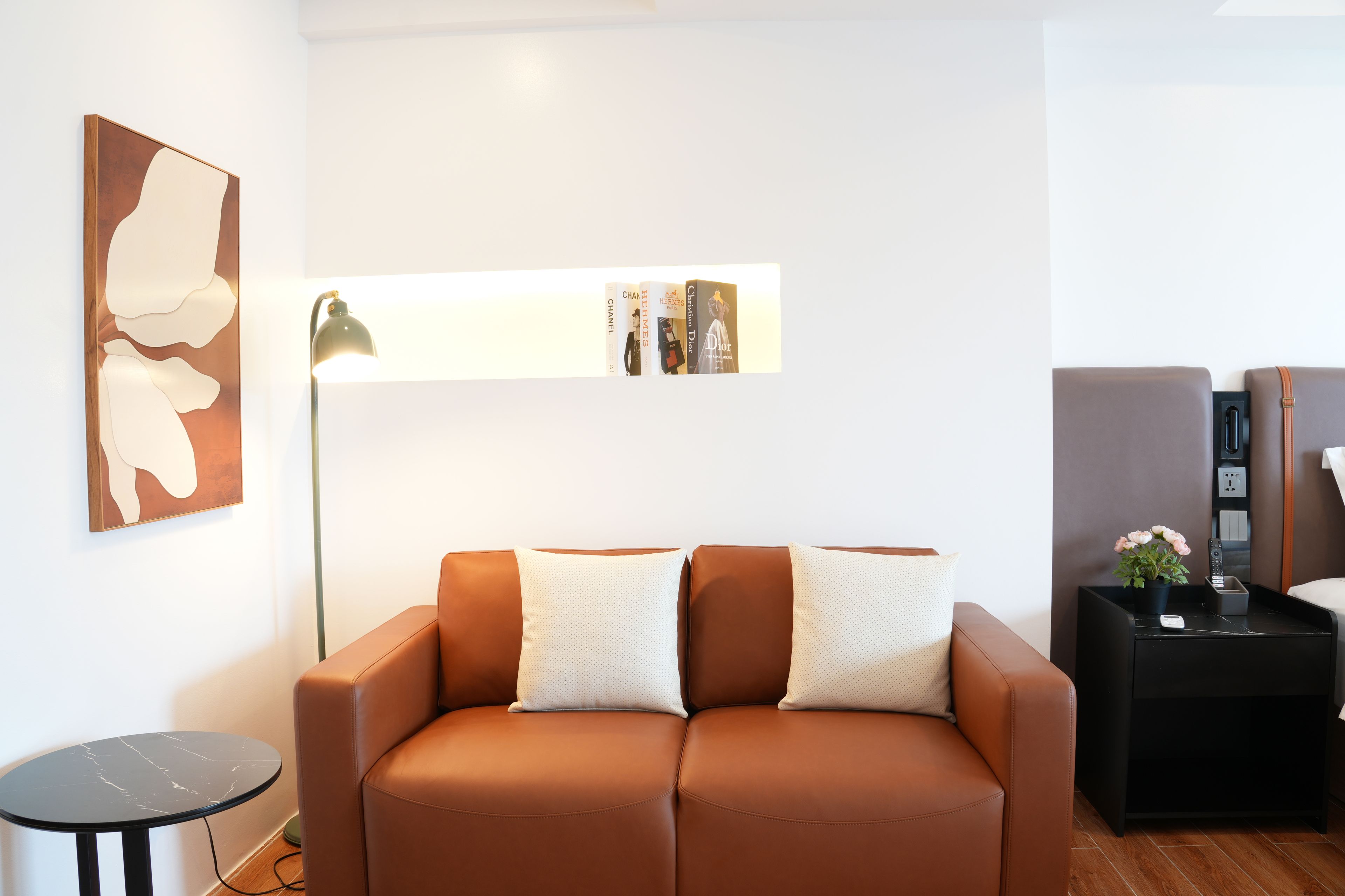 Una sala de estar moderna con un sofá de cuero marrón, una mesita de noche con un control remoto y una planta, y un estante montado en la pared con libros.
