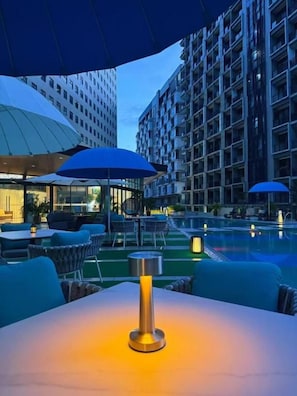 Un área de estar al aire libre con mesas y sombrillas, con vista a una piscina y edificios cercanos.