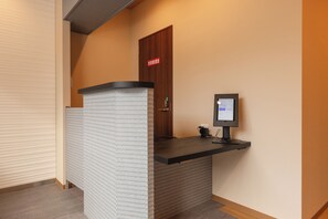 En receptionsdisk med en computerskærm, en selvbetjeningsautomat til check-in og en dør med et skilt.