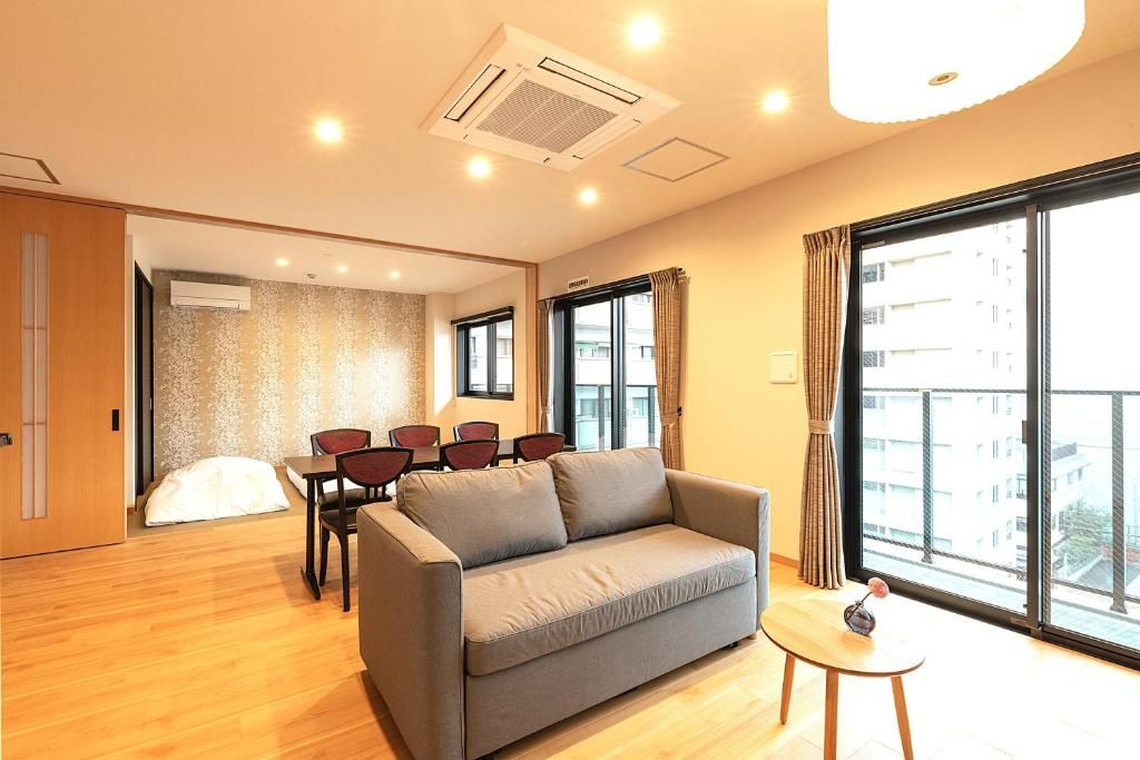 A modern hotel room with a sofa, a dining table with chairs, and a large window offering a view of the city.