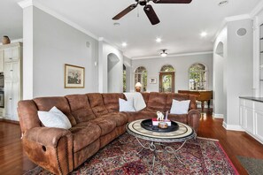 A spacious living room with a brown leather sofa, a round coffee table, and a patterned rug. There is a ceiling fan, a piano, and a dining area visible in the background. - Whispering Pines Manor - Luxury Lake Retreat (Northport)