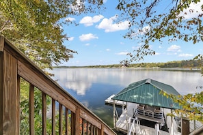 A wooden deck overlooking a calm lake with a dock and a covered boat house. - Whispering Pines Manor - Luxury Lake Retreat (Northport)