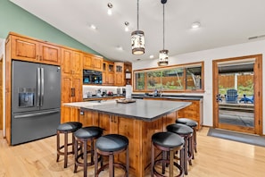 A modern kitchen with wooden cabinets, a stainless steel refrigerator, a large island with bar stools, and a view of the backyard through a sliding glass door. - Mnt Elden Base Camp - 3BD/2BTH - Sleeps 8 - Trails (Flagstaff)