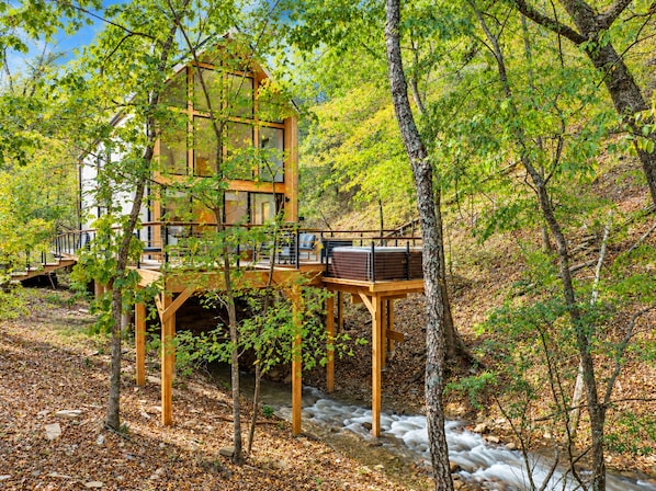 A wooden cabin with a glass facade, situated in a forested area with a stream flowing nearby.