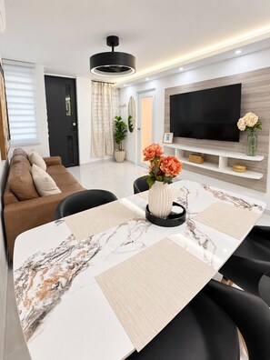 A modern living room with a sofa, a dining table with placemats, a TV on the wall, and a potted plant.