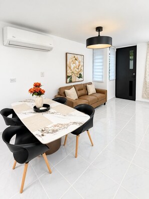 A modern living room with a dining area, featuring a sofa, a dining table with chairs, a black pendant light, and a large floral painting on the wall.