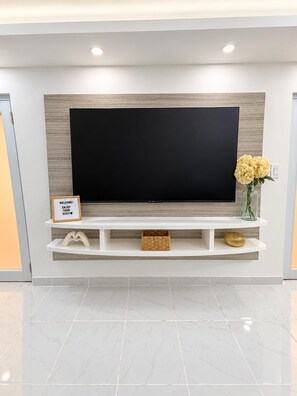 A modern living room with a large flat-screen TV mounted on a wooden panel wall, a white entertainment unit below, and a vase with flowers on the right. - Carolina Luxe 2BR Escape Near SJU (Carolina)