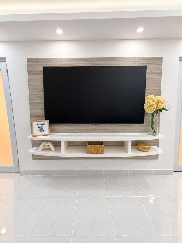 A modern living room with a large flat-screen TV mounted on a wooden panel wall, a white entertainment unit below, and a vase with flowers on the right.