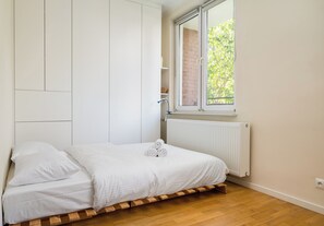 A bedroom with a bed, white walls, a window, and a radiator. - Park Moczydło Cozy Studio (Warszawa)