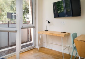 A room with a wooden desk, a chair, a TV mounted on the wall, and a window with a view of trees. - Park Moczydło Cozy Studio (Warszawa)
