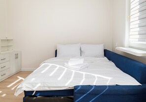 Un letto rifatto con lenzuola bianche e un asciugamano piegato, una testiera imbottita blu e un comò bianco con cassetti.