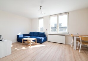 A modern living room with a blue sofa, a wooden coffee table, a white radiator, and a window with a view of buildings. - Ochota Green Spot Studio (Warszawa)