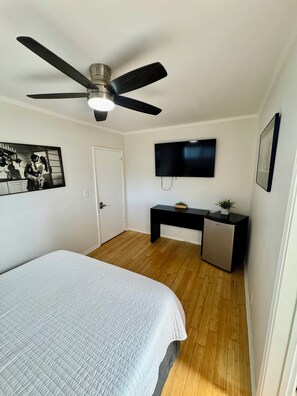 A bedroom with a bed, a ceiling fan, a TV, a desk, and a small fridge. - Charming bedroom in private home (Hawthorne)