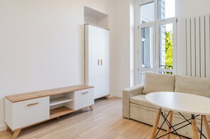 A modern living room with a sofa, a round table, a white cabinet, and a window with a view of greenery. - Urban Nest Modern Studio (Katowice)