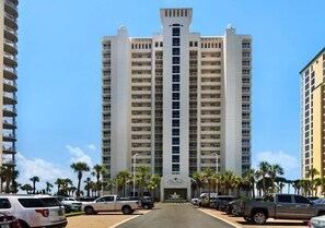 A multi-story hotel with a central entrance, surrounded by parked vehicles and palm trees. - Gulf Front Views •Pool •Hot Tub | The Pearl 1201 (Gulf Breeze)