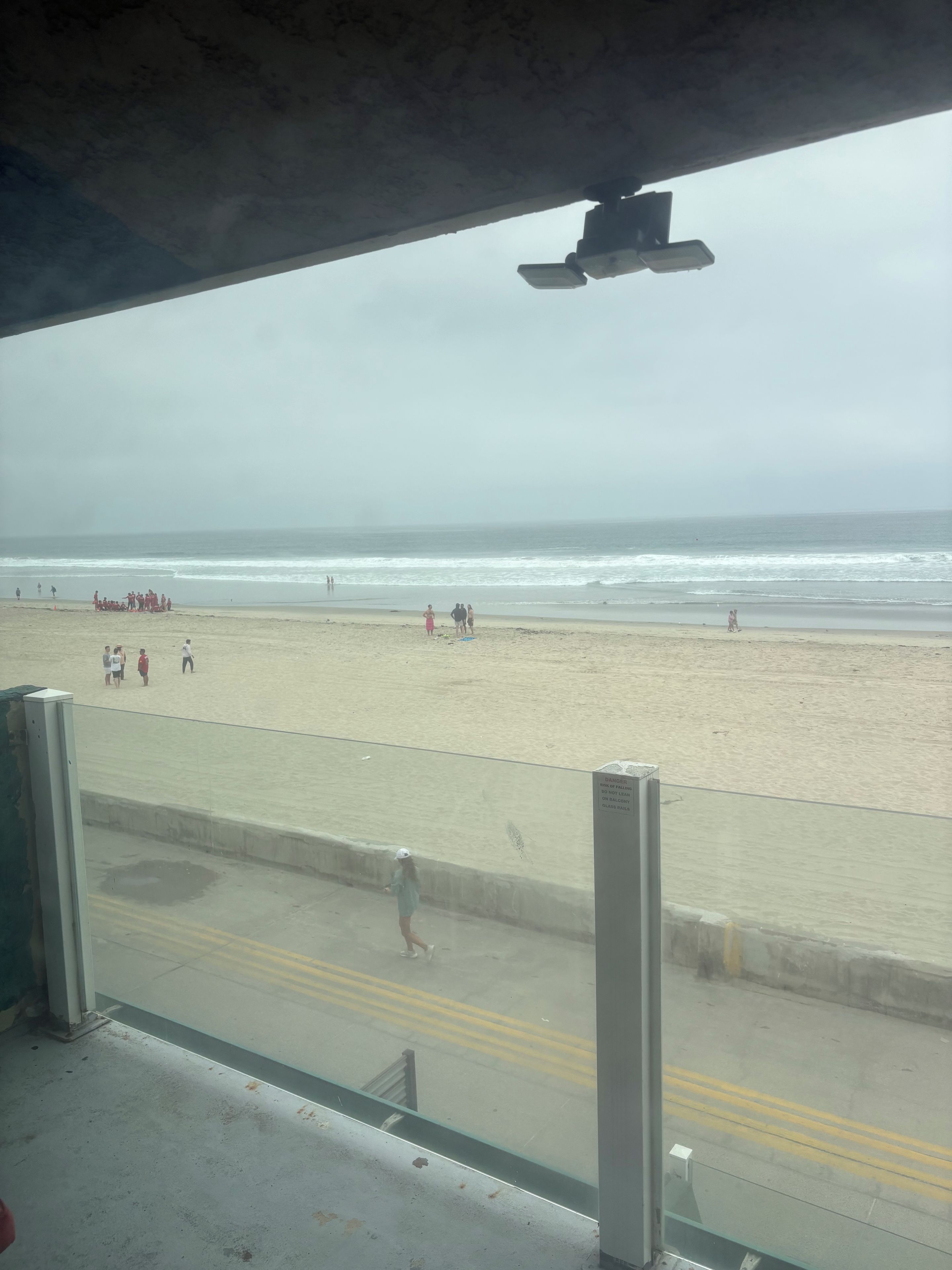Vista a la playa desde un espacio techado con piso de concreto y una barrera de vidrio.