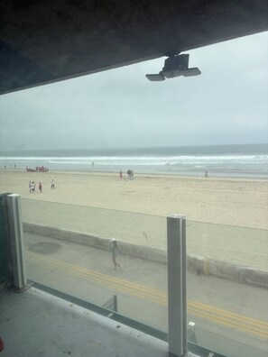 Vista a la playa desde un espacio techado con piso de concreto y una barrera de vidrio.