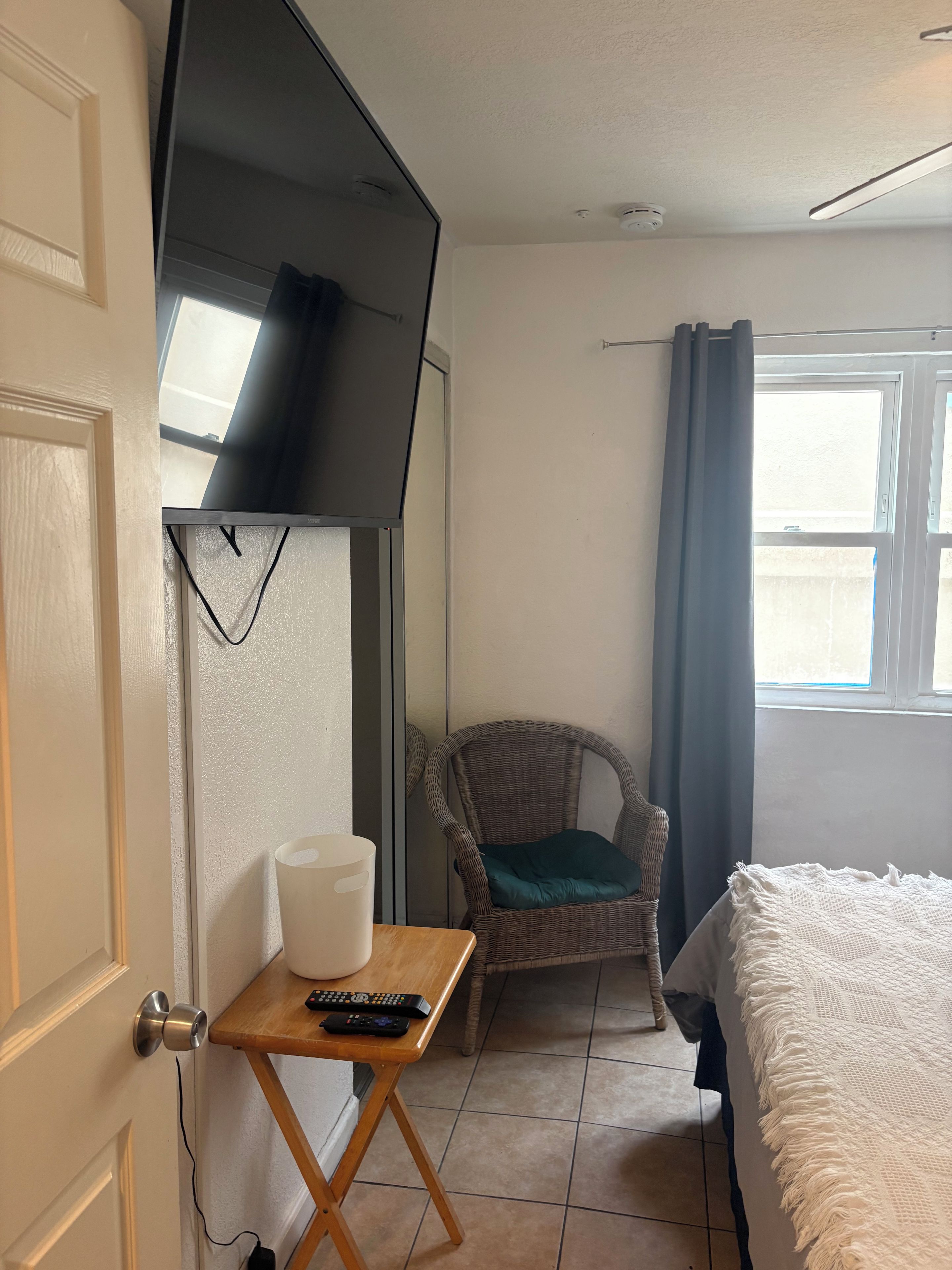 Una habitación con una cama, una silla, una mesita con un control remoto y un televisor montado en la pared.