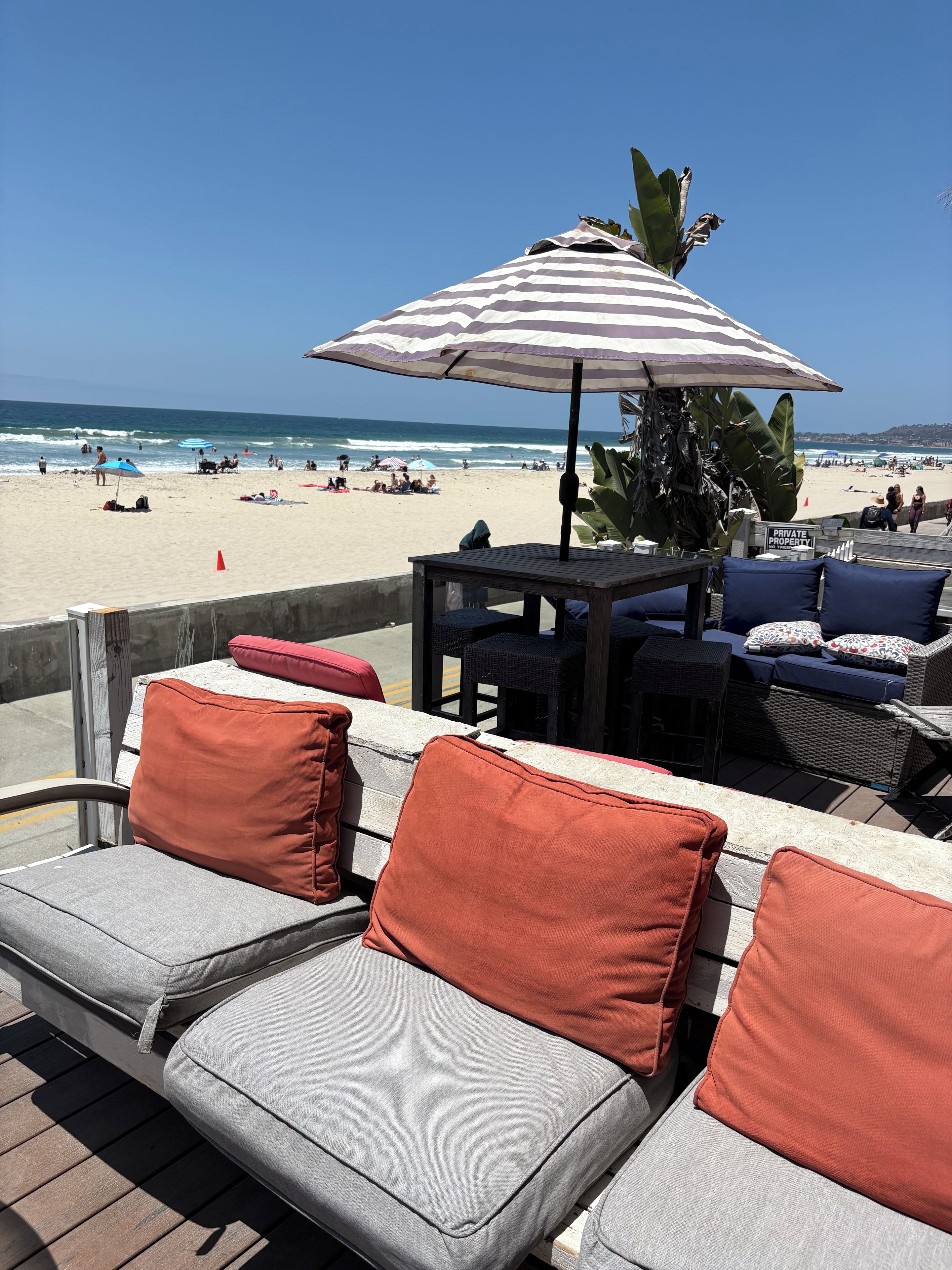 Un patio frente a la playa con mobiliario exterior, una sombrilla a rayas y vista al mar.
