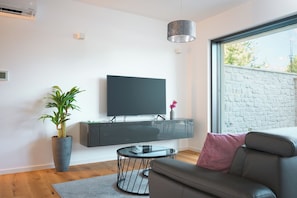 Ein modernes Wohnzimmer mit einem wandmontierten Flachbildfernseher, einem runden Glastisch, einem grauen Sofa, einer Zimmerpflanze und einem großen Fenster mit Blick auf Bäume.