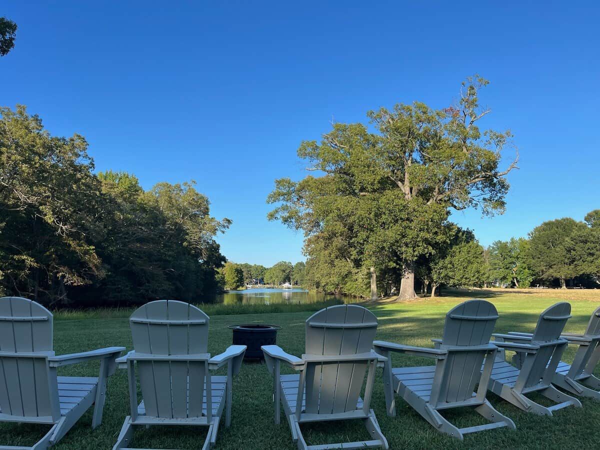 A row of Adirondack chairs facing a lake with trees in the background.