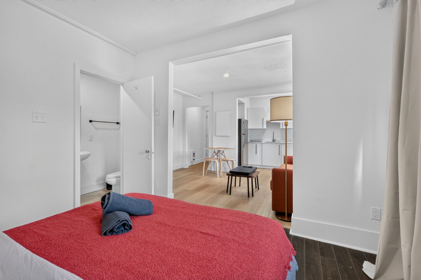 A hotel room with a bed, a small table, a chair, a desk, and a kitchen area visible through an open door.