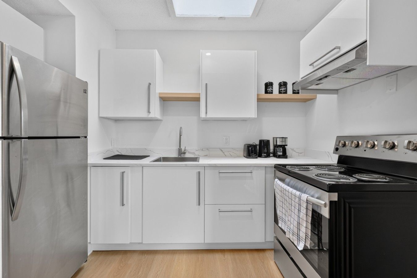 A modern kitchen with white cabinets, a stainless steel refrigerator, a sink, a coffee maker, and a stove.