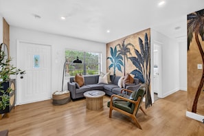 A modern living room with a grey sectional sofa, a round wooden coffee table, a green armchair, and a large tropical-themed wall mural. - Casa Palma Heated Pool Hot Tub Games (Fort Lauderdale)