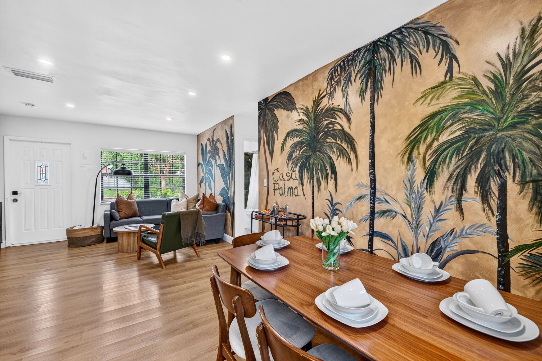 A dining area with a wooden table set for four, a sofa, and a large wall mural of palm trees.