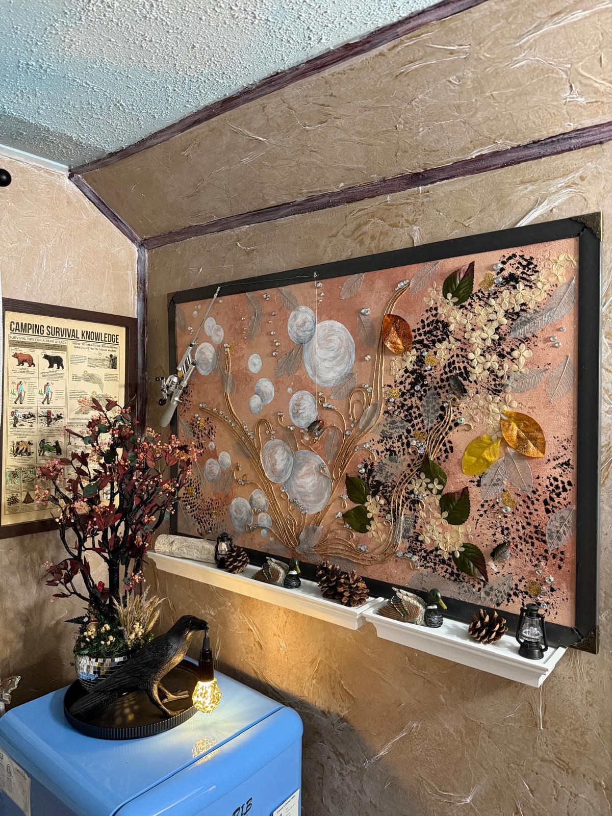A wall with a framed artwork depicting a tree with leaves and flowers, a shelf with decorative items, and a framed poster on the wall.