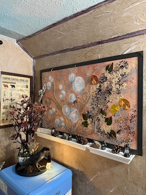 A wall with a framed artwork depicting a tree with leaves and flowers, a shelf with decorative items, and a framed poster on the wall.