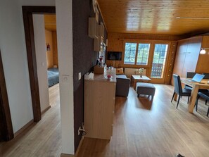 Ein geräumiges Zimmer mit Holzbalkendecke, einem Essbereich mit Tisch und Stühlen, einer Küchenzeile mit Arbeitsplatte und Schränken sowie einem großen Fenster mit Blick ins Freie.