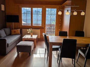 Una stanza accogliente con pavimento in legno, un divano, un tavolo da pranzo con sedie e ampie finestre vista alberi.