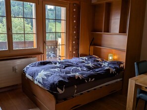 Un letto di legno con copripiumino a motivi fogliari in una stanza con pareti in legno e un'ampia finestra.