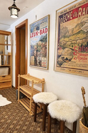 Ein Zimmer mit einer hölzernen Bank und zwei mit Fell bezogenen Hocker, einem gemusterten Teppich und einer Wand mit alten Plakaten.