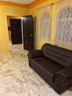 A room with a brown leather sofa, a door leading to another room, and a window with a geometric pattern. - Beautiful 4-bedroom apartment with AC in Saint Ann's Bay (Saint Ann's Bay)