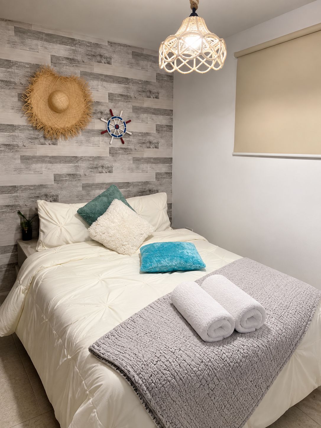 Une chambre à coucher avec un lit, des oreillers, une couverture, un chapeau accroché au mur, une décoration représentant un gouvernail de bateau et un luminaire suspendu.