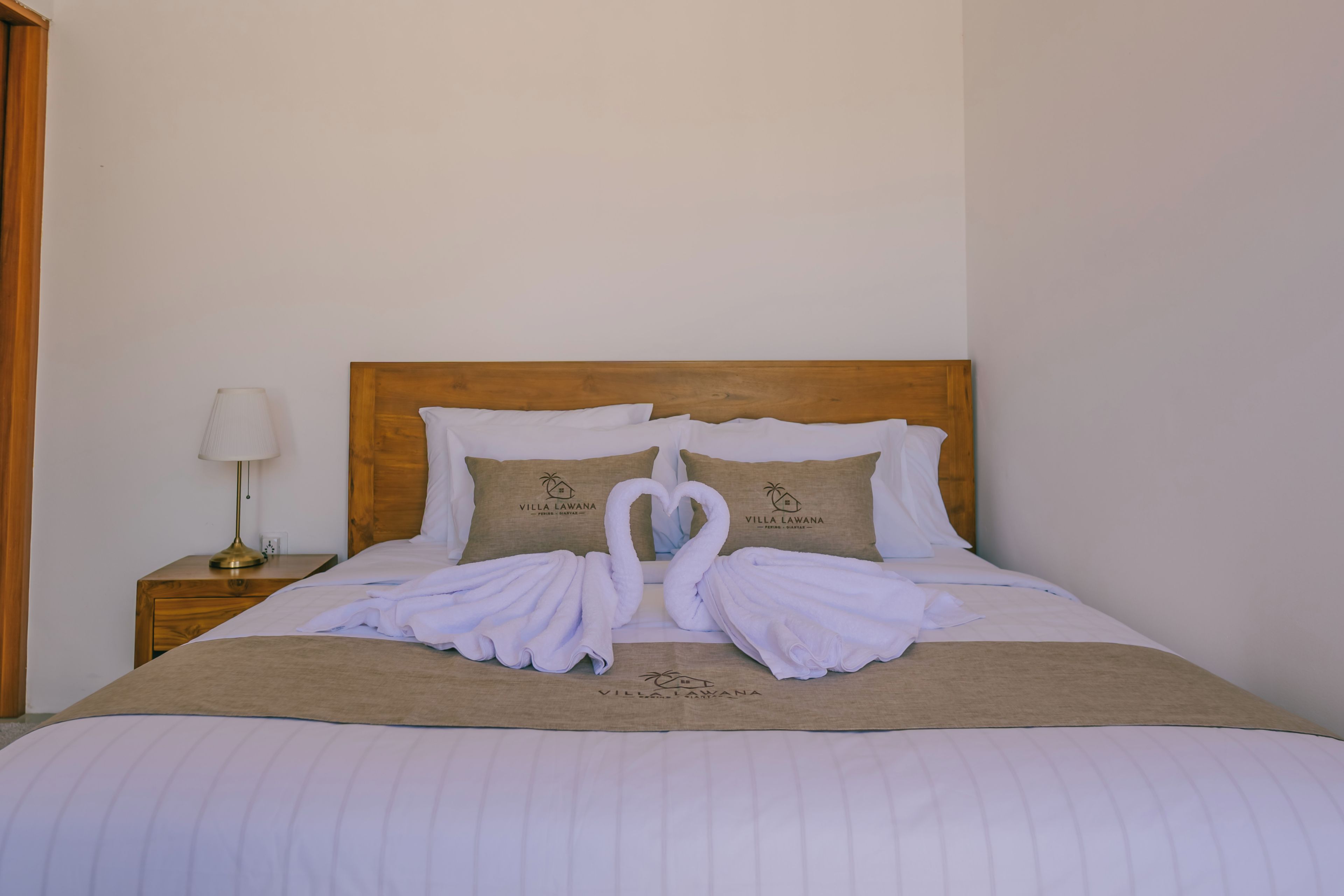A neatly made bed with a wooden headboard, two pillows with the Villa Lawana logo, and a towel arrangement in the shape of a swan.