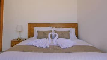 A neatly made bed with a wooden headboard, two pillows with the Villa Lawana logo, and a towel arrangement in the shape of a swan.