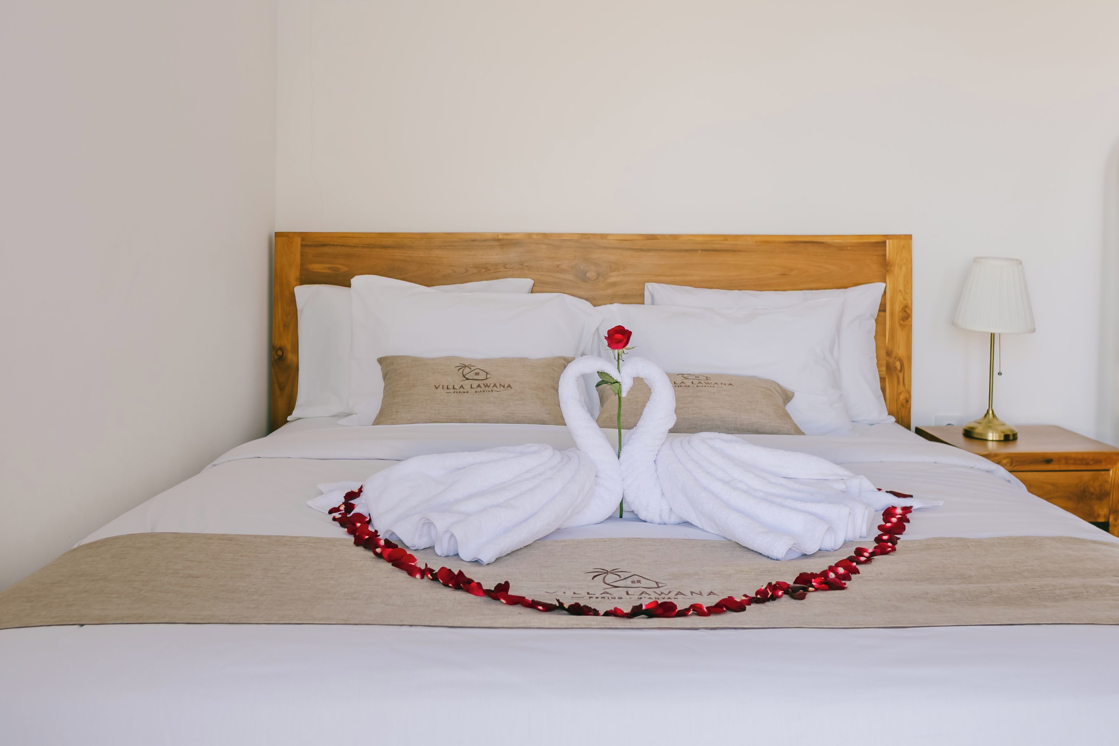 Una cama bien tendida con una decoración de toallas con forma de cisne y pétalos de rosa, cabecera de madera, ropa de cama blanca y una lámpara de noche.