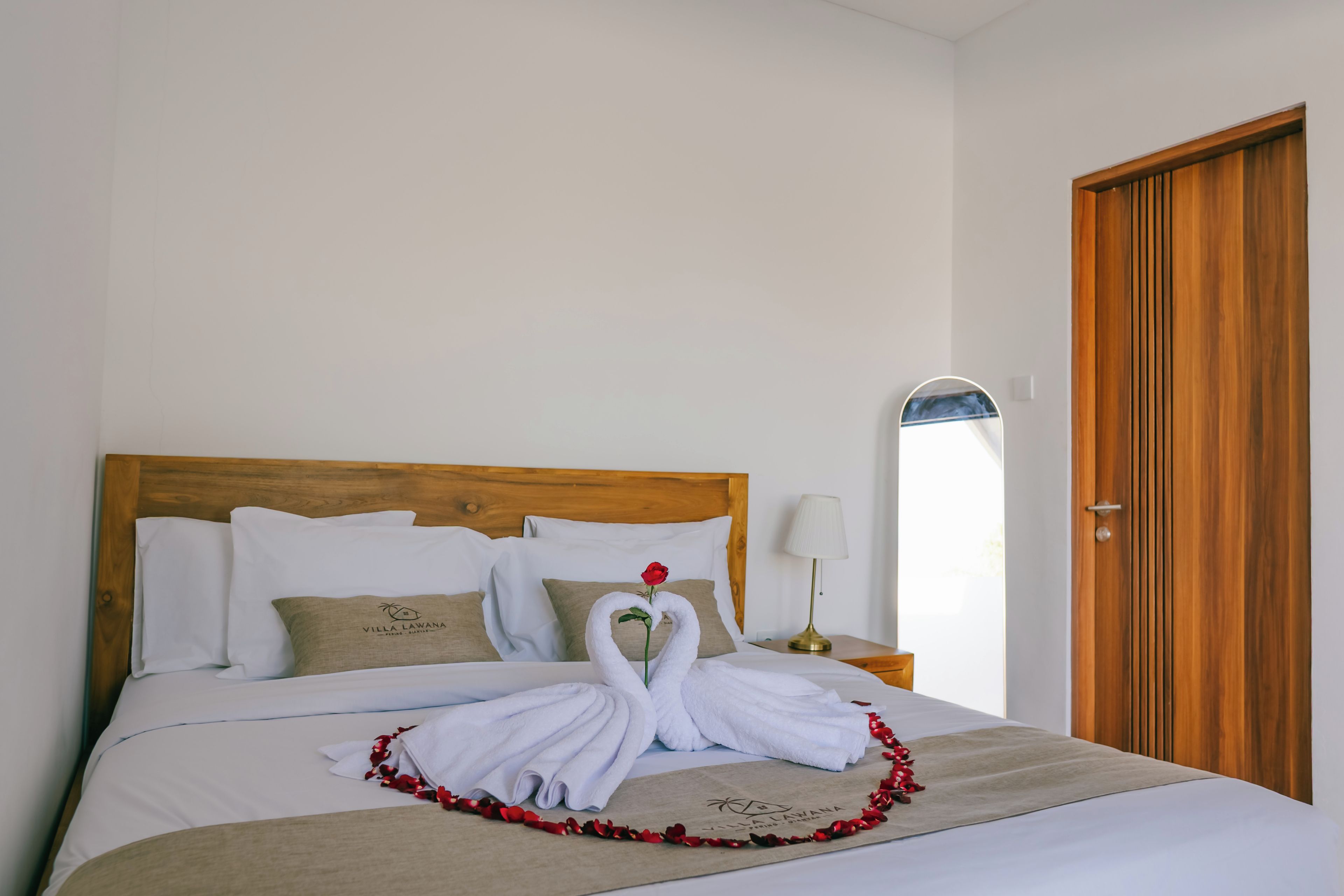 A hotel room with a neatly made bed, a wooden headboard, a nightstand with a lamp, and a wooden door.